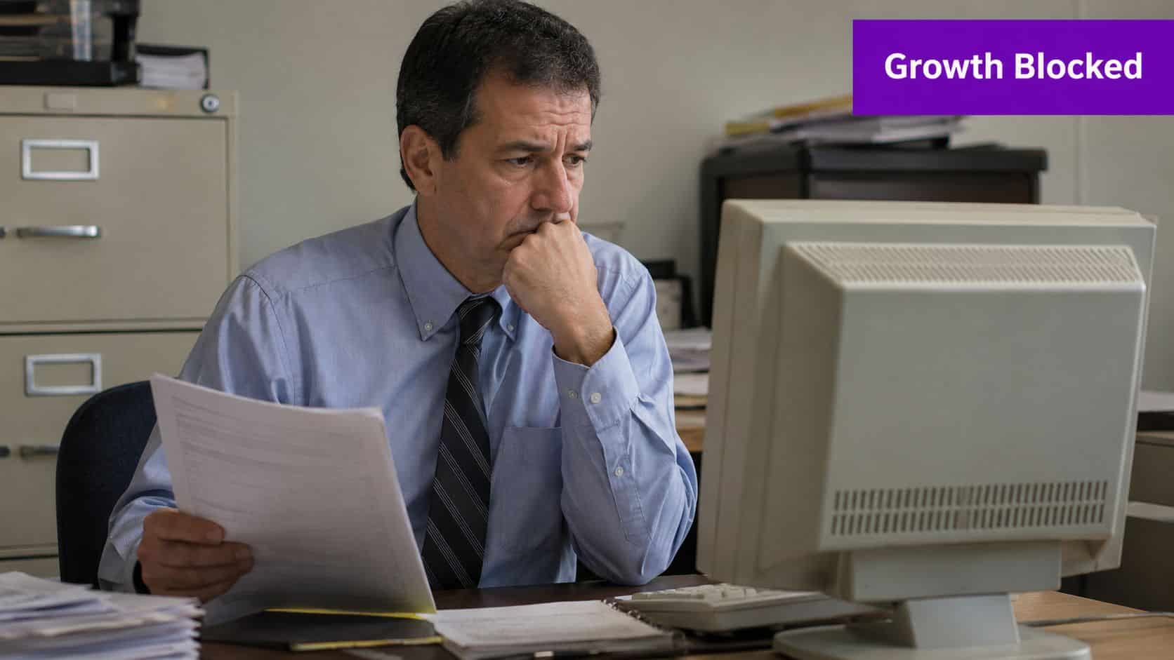 A concerned office worker looks at a computer screen while holding a document at his desk.