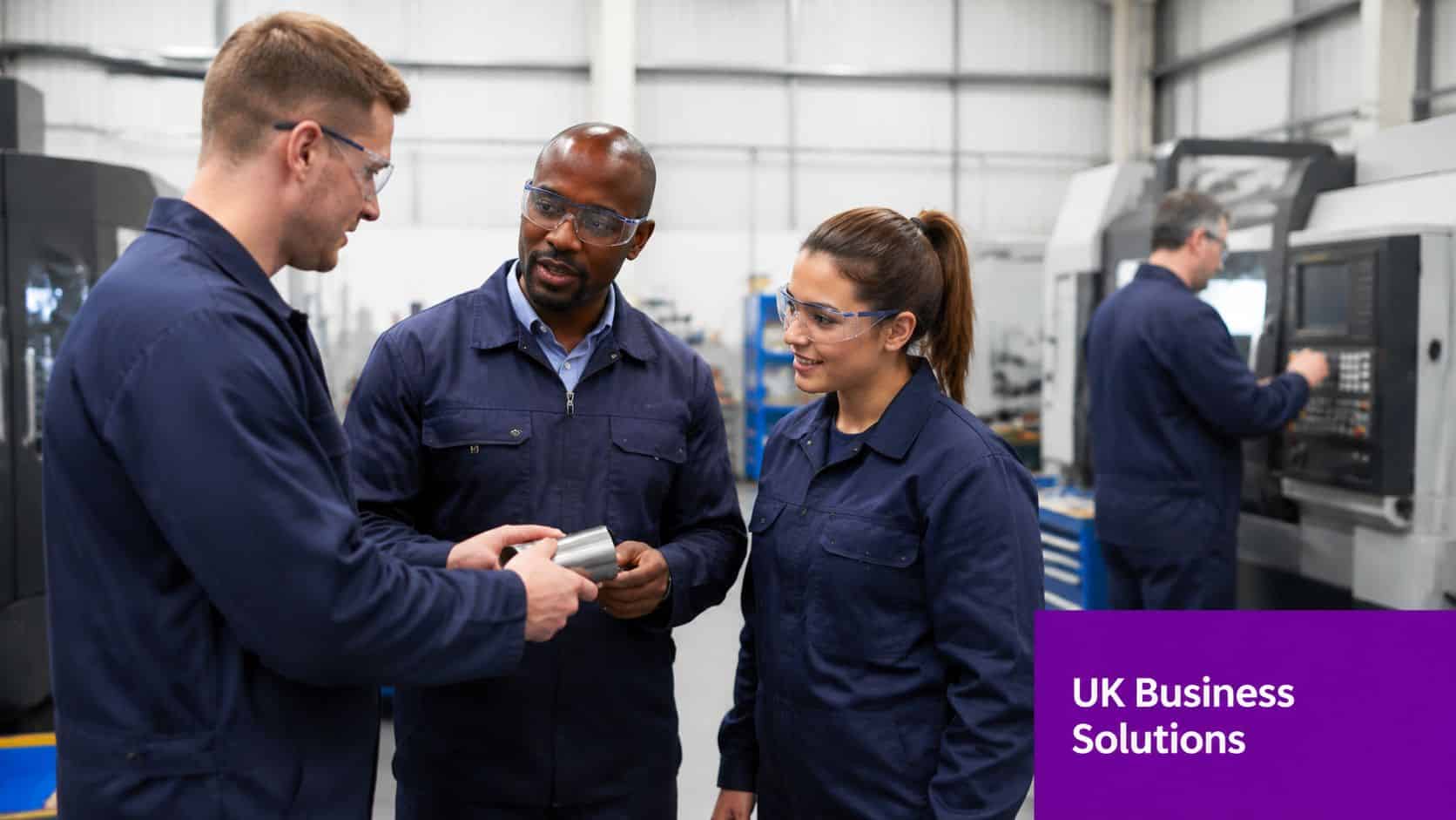Three factory workers discussing a metal part while another colleague operates machinery in the background.